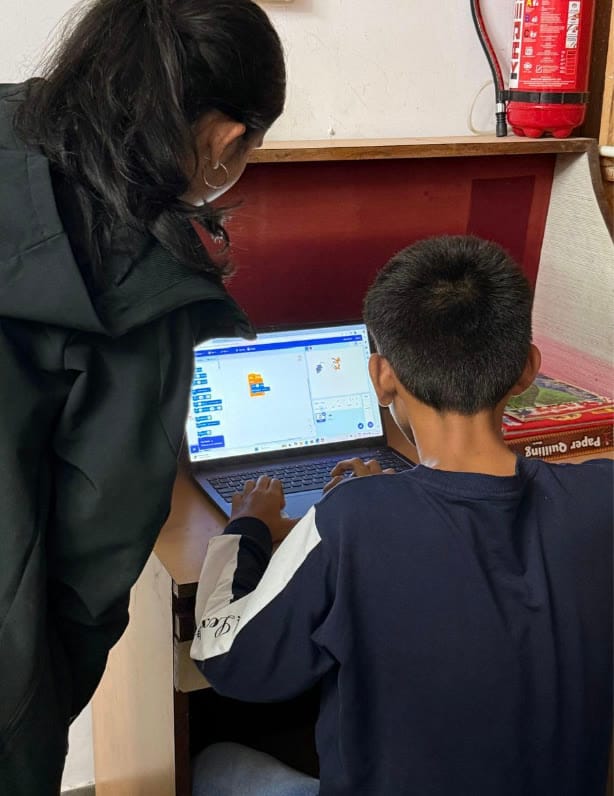 A student's hands on a keyboard, Scratch on screen, other children watching.